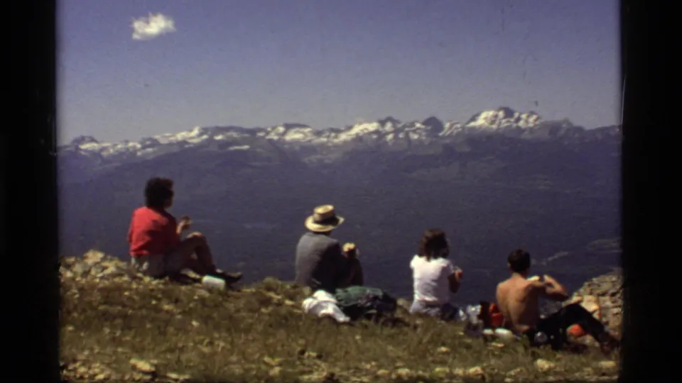 1977 8mm frame at Sapphire Lake, Montana, with hikers resting on grass and rocks while viewing a hazy snow-capped mountain range.