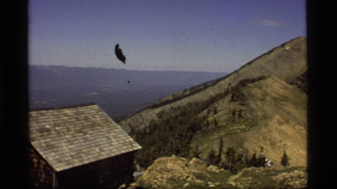Mountain ridge with cabin roof, distant lake, blue sky, and small hiker below.