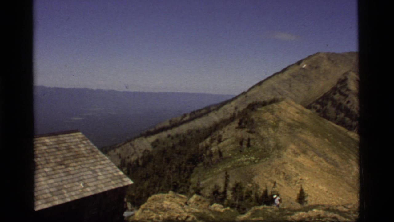 Sapphire Lake, Montana home-movie frame showing a cabin roof, broad ridge, distant water, and a small walker near the lower slope.