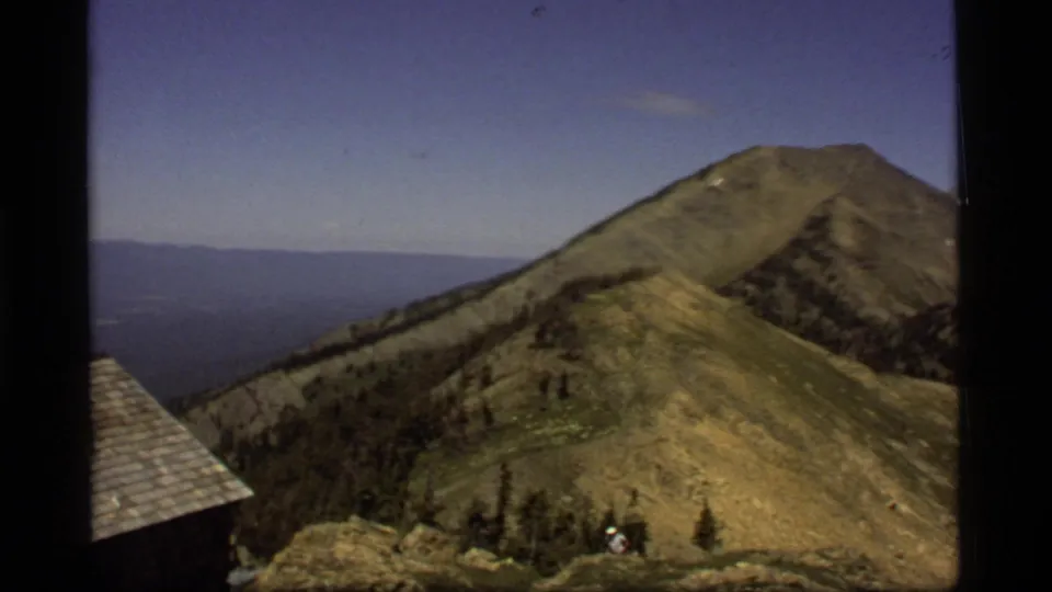 1977 8mm frame of a Montana ridge and peak, with a partial cabin roof at left and a tiny person near rocks and trees below.