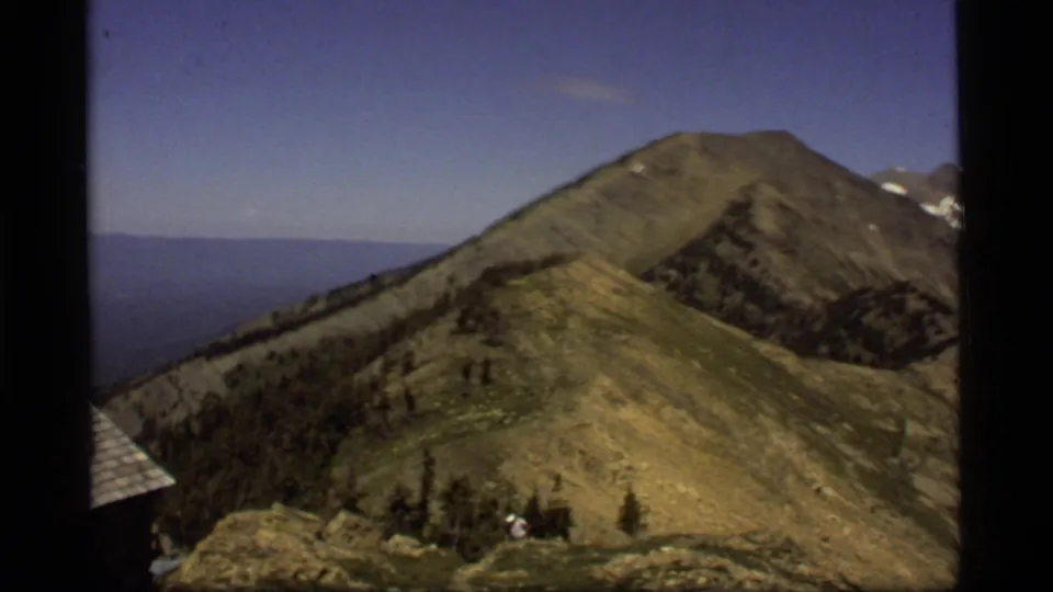 Vintage 8mm view of a Sapphire Lake, Montana ridge with a cabin edge, distant lake, and a small person near the rocky foreground.