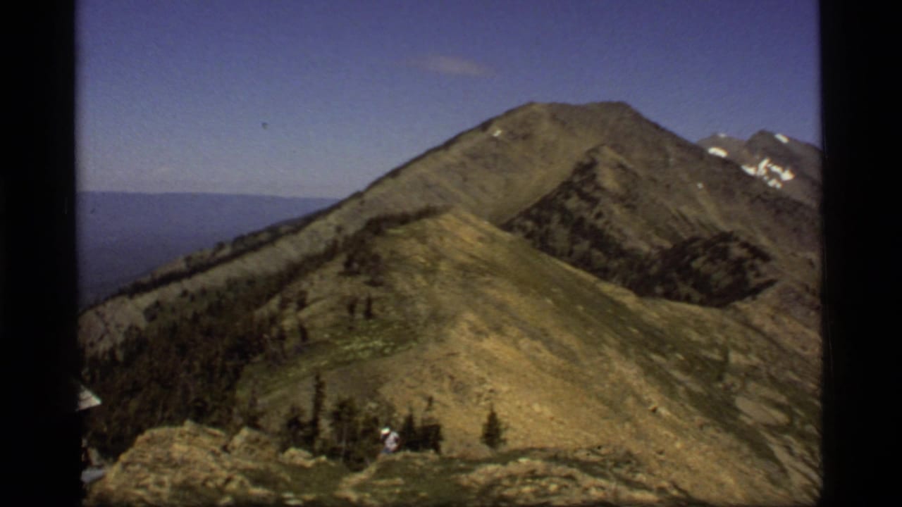 1977 8mm Montana home-movie frame of a small hiker on a rocky ridge near grassy slopes and distant snow-capped peaks.