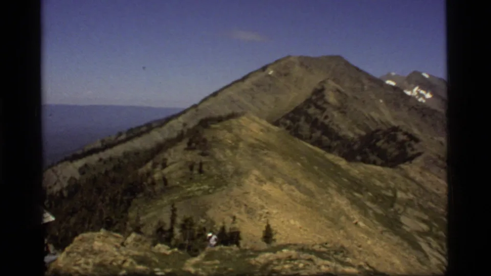 1977 8mm Montana home-movie frame of a small hiker on a rocky ridge near grassy slopes and distant snow-capped peaks.