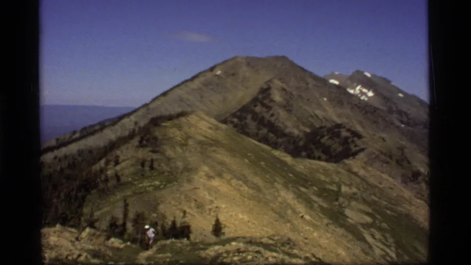 1977 8mm frame from Montana showing a hiker on a rocky ridge with green-brown slopes and snowy peaks beyond.