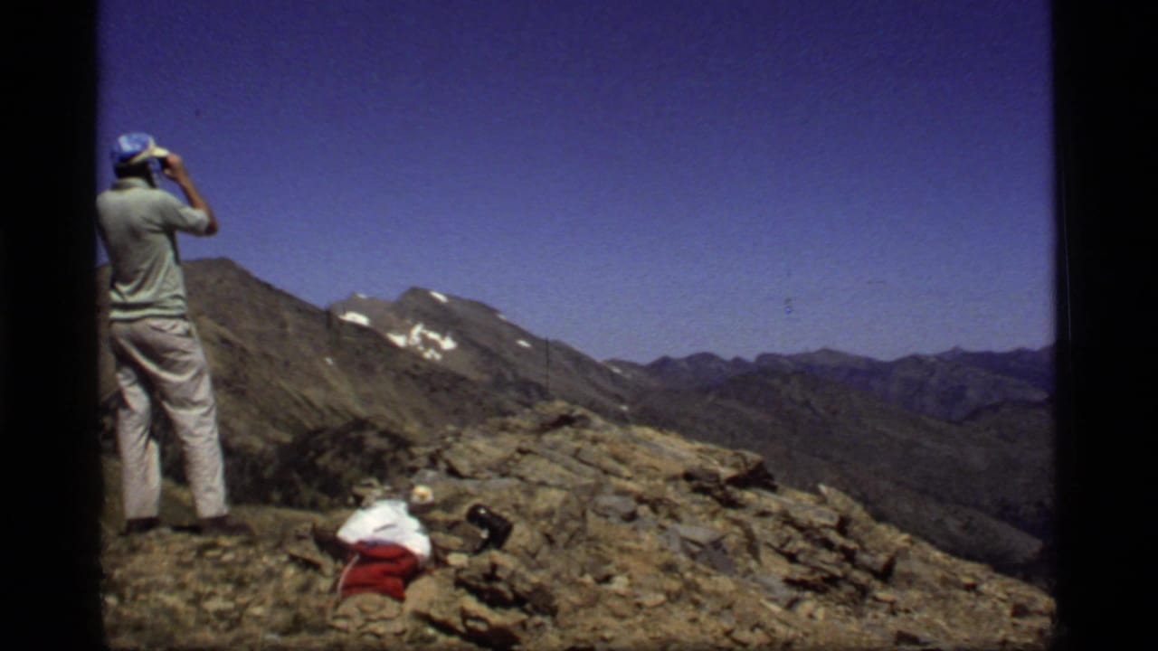 1977 Montana home-movie frame showing a man in light clothes using binoculars on a rocky overlook with distant peaks behind him.