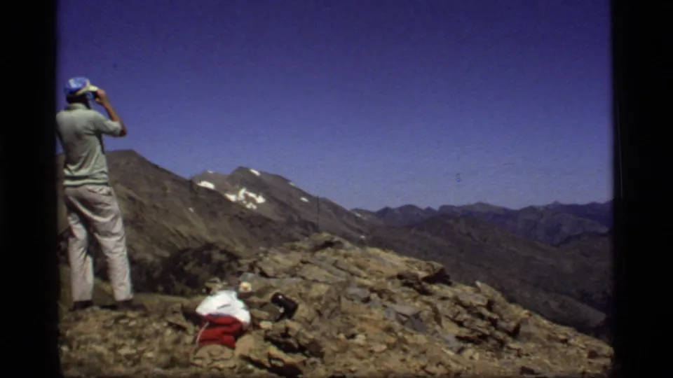 1977 Montana home-movie frame showing a man in light clothes using binoculars on a rocky overlook with distant peaks behind him.