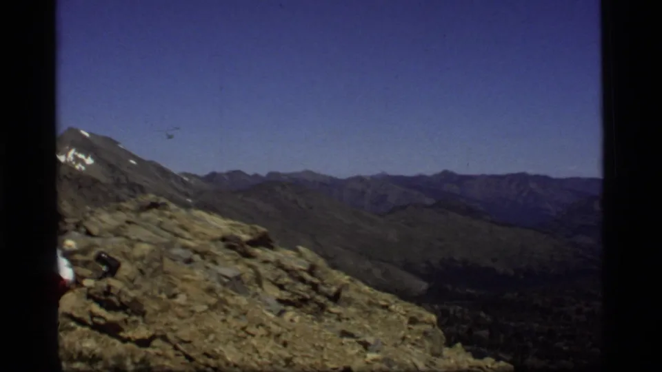 1977 Montana home-movie view across rocky terrain toward layered mountain ridges, with the hiker's pack partly visible at frame left.
