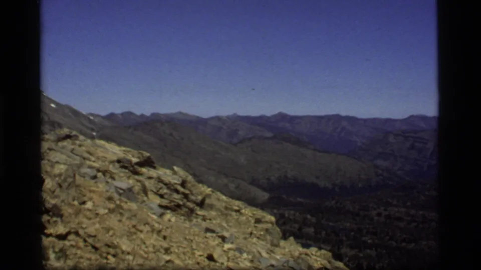 1977 Montana home-movie frame focused on rocky high ground, forested valley terrain, and layered mountain ridges under a clear sky.