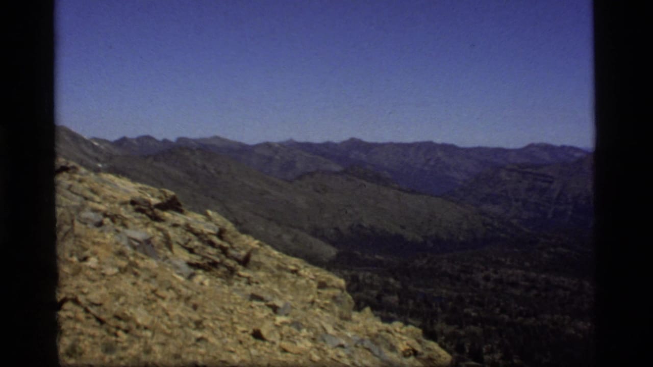 1977 8mm view from Sapphire Lake, Montana, looking across tan rocks, forested valley, and layered mountain ridges.