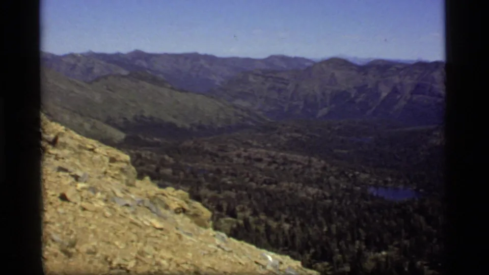 1977 home-movie frame from Sapphire Lake, Montana, with sunlit rocks, a forested valley, distant ridges, and a small lake.