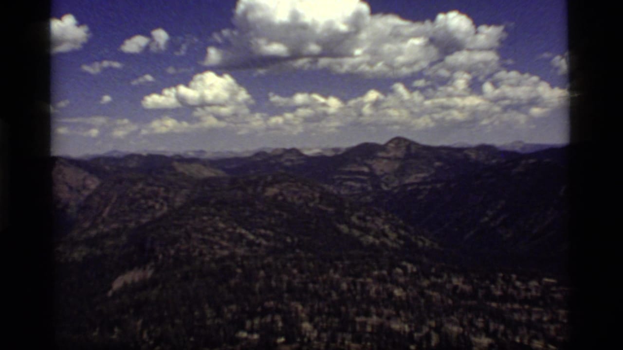 1977 8mm Sapphire Lake, Montana vista with layered mountain ridges, dark forested slopes, and bright cumulus clouds overhead.