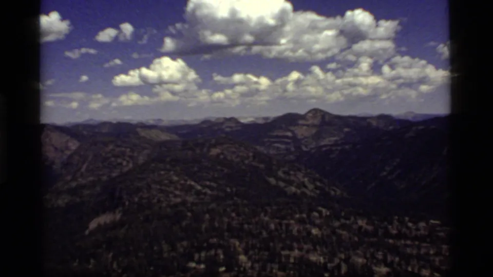 1977 8mm Sapphire Lake, Montana vista with layered mountain ridges, dark forested slopes, and bright cumulus clouds overhead.