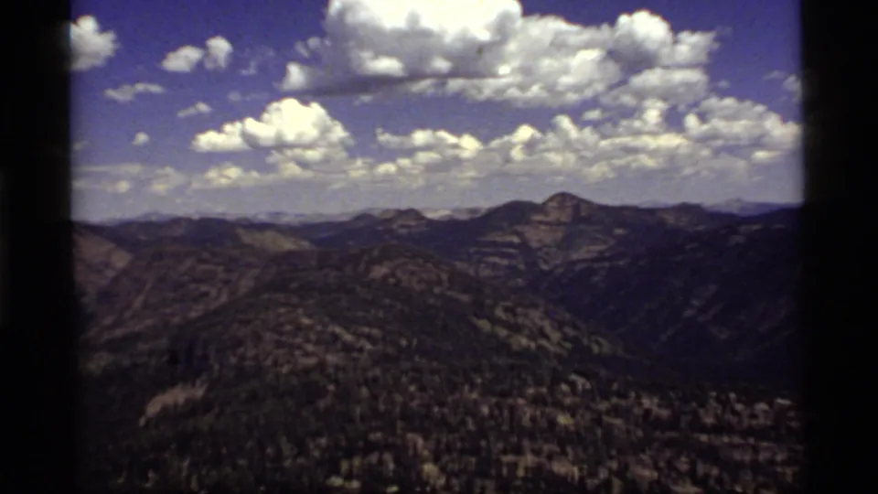 1977 8mm mountain panorama near Sapphire Lake, Montana, with rolling foothills, wooded slopes, and clustered white clouds.