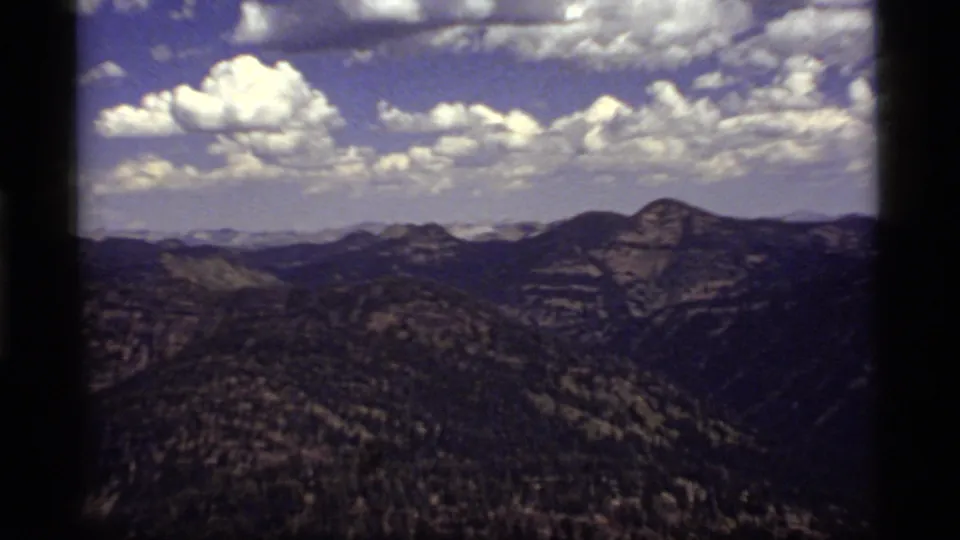 1977 8mm Sapphire Lake, Montana landscape looking across dark forested valleys toward rounded mountain ridges and bright clouds.