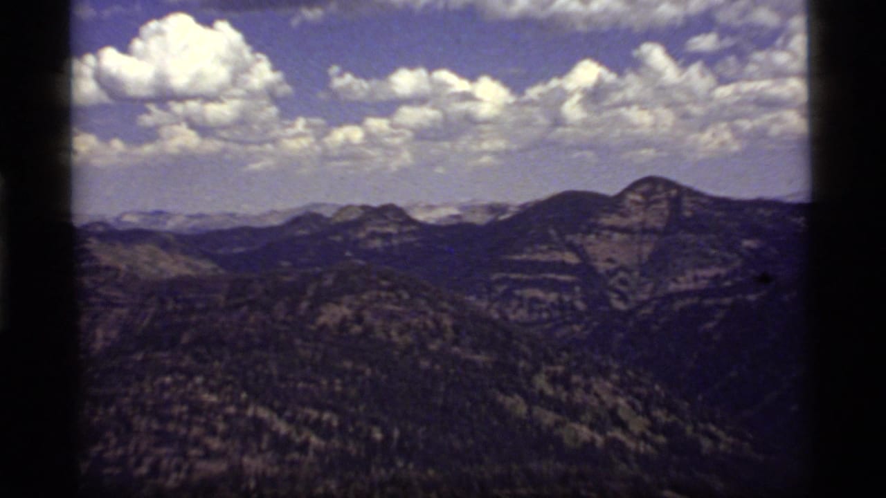 1977 8mm view near Sapphire Lake, Montana, showing layered forested ridges beneath bright scattered clouds.