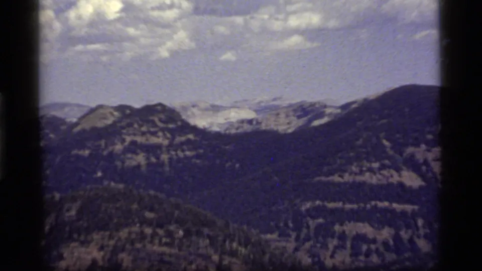 1977 Montana home-movie frame showing pale distant slopes beyond dark forested ridges near Sapphire Lake.