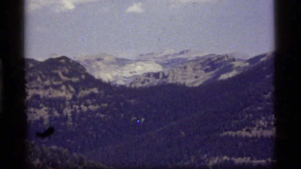 1977 8mm Montana home-movie frame showing distant rugged mountains, forested slopes, and a blue-tinted sky.