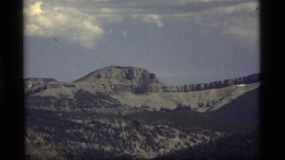 1977 8mm landscape frame showing a broad Montana mountain formation, wooded slopes, and a lightly clouded sky.