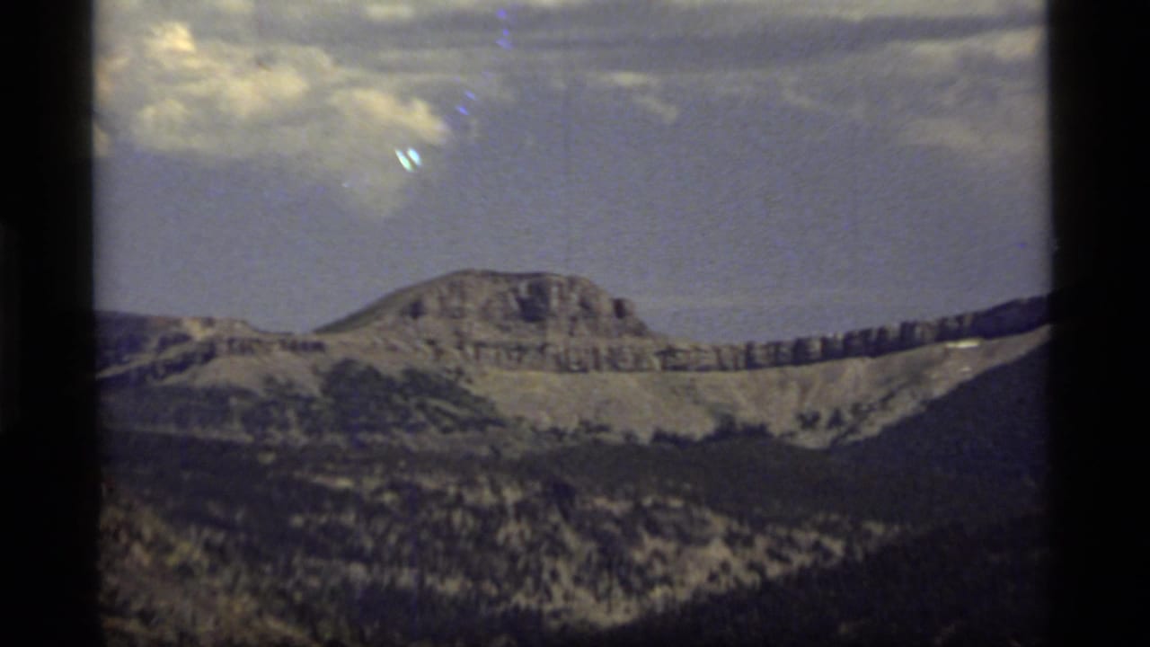 1977 Montana 8mm frame showing a distant rocky mountain ridge, lower forested slopes, light snow patches, and a bright clouded sky.