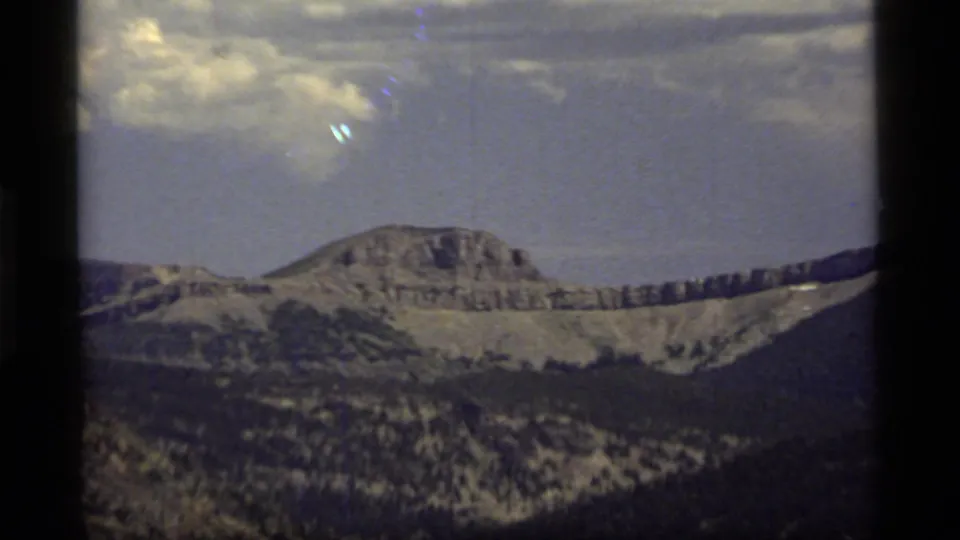 1977 Montana 8mm frame showing a distant rocky mountain ridge, lower forested slopes, light snow patches, and a bright clouded sky.
