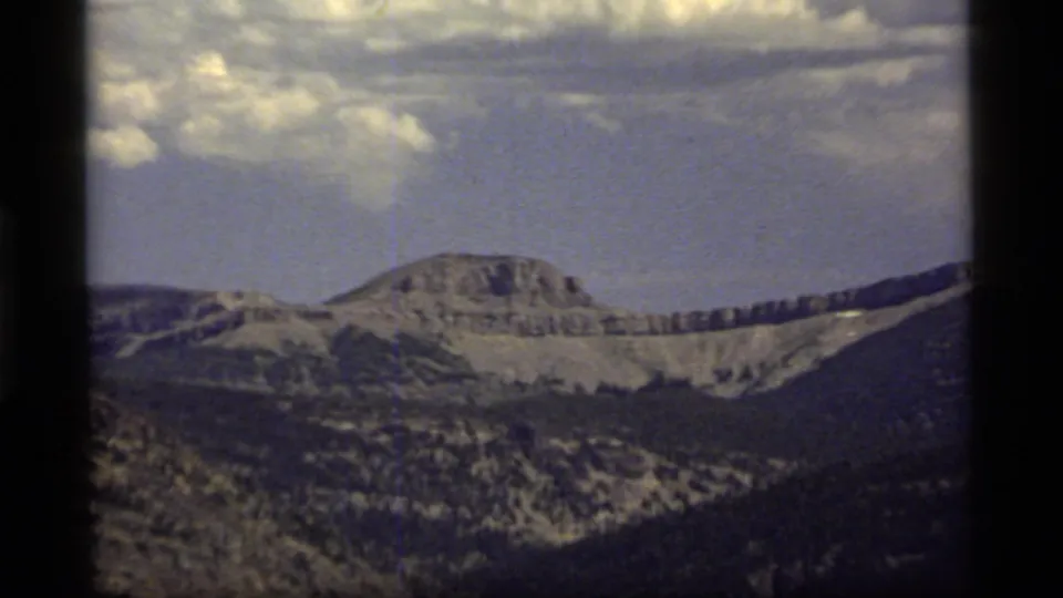 1977 Sapphire Lake, Montana footage frame with a broad ridgeline, layered hills, forested slopes, small snow patches, and visible 8mm frame edges.