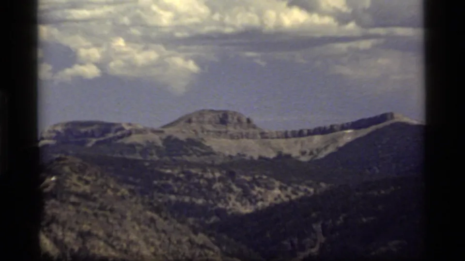 1977 Montana home-movie frame showing a rocky mountain ridge above a forested valley, with soft focus, muted color, and dark film-frame borders.
