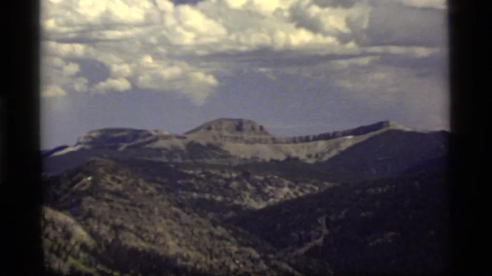 1977 Sapphire Lake, Montana 8mm frame showing a distant mountain ridge, forested lower slopes, patchy snow, bright clouds, and heavy frame-edge shadow.