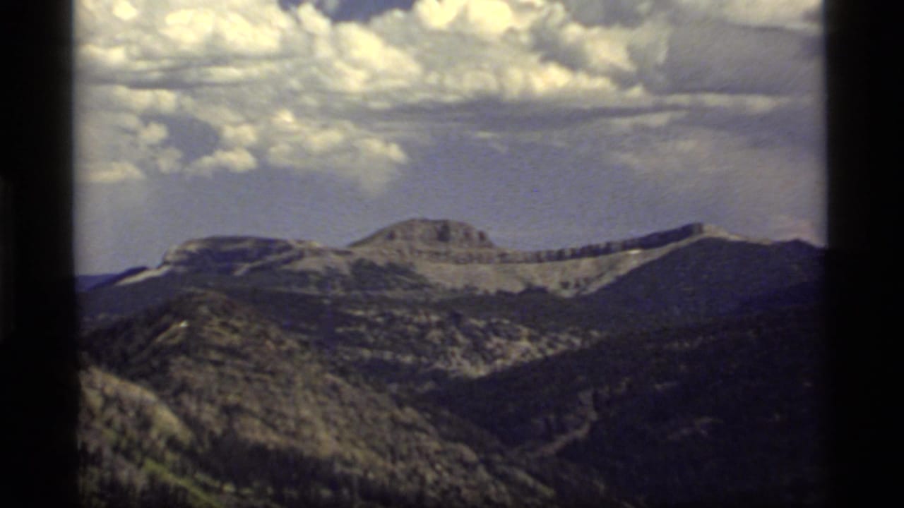 1977 8mm home-movie view of mountain ridges near Sapphire Lake, Montana, with forested slopes and large clouds above.