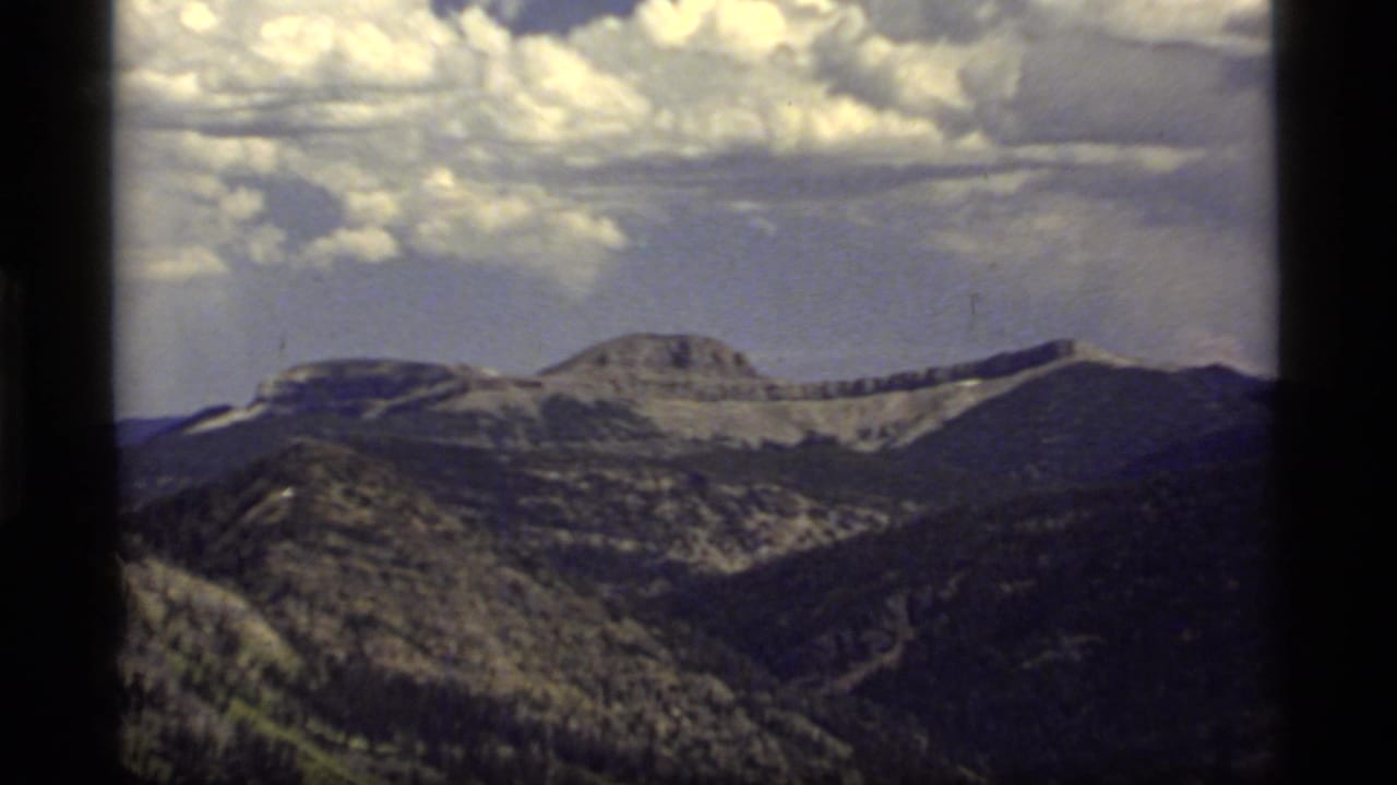 Vintage 8mm frame of Sapphire Lake area mountain terrain, with distant rocky ridges, wooded slopes, and a dramatic cloud bank.