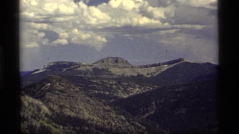 Vintage 8mm frame of Sapphire Lake area mountain terrain, with distant rocky ridges, wooded slopes, and a dramatic cloud bank.