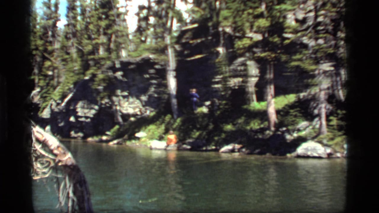 1977 8mm frame from Sapphire Lake, Montana, showing two men separated by the shoreline, trees, rocks, and green water.