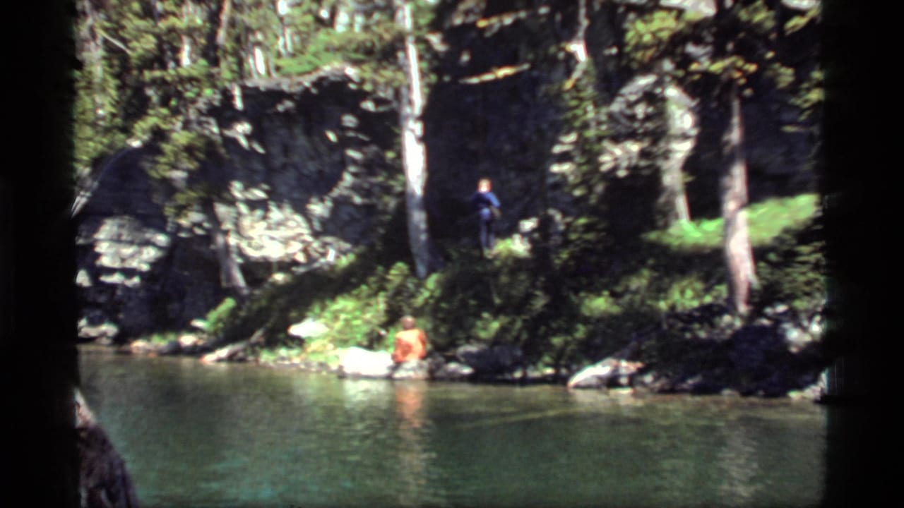 Sapphire Lake, Montana home-movie frame with a seated man by the water and a standing man on a rocky forest bank.