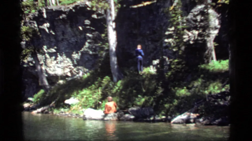 1977 Sapphire Lake frame showing a man seated near the water and another standing on the shaded bank above.