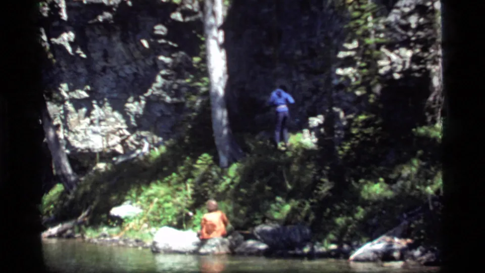 Home-movie frame from Sapphire Lake, Montana, with two men at a shaded rocky shoreline under tall trees.