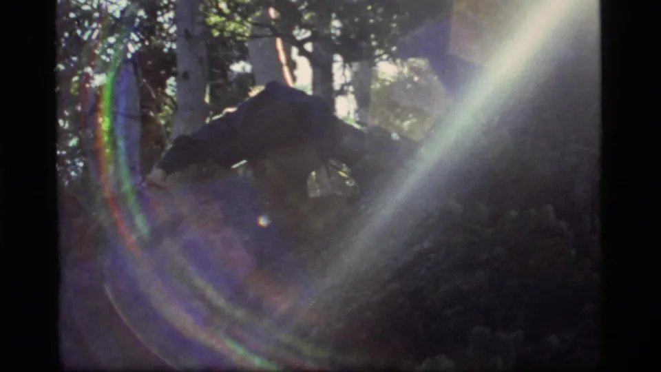 A late-1970s 8mm frame shows a person low to the ground in the Sapphire Lake forest, partly obscured by bright rainbow flare.
