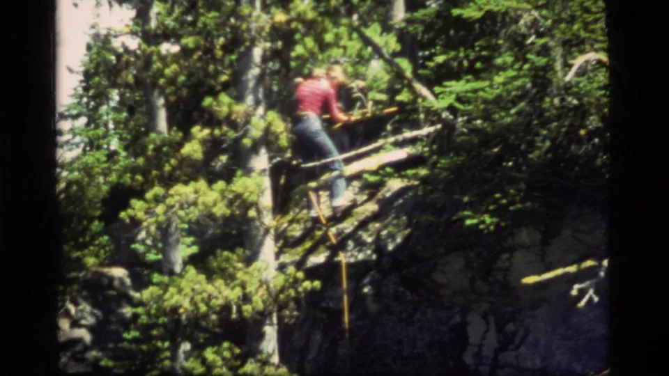 A 1977 home-movie frame shows the person leaning over the rough bridge structure in a rocky Montana forest setting.