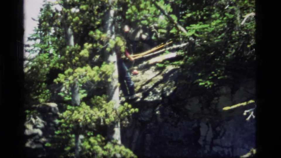 1977 Sapphire Lake, Montana home-movie frame of a distant worker on a steep evergreen-covered rock slope.