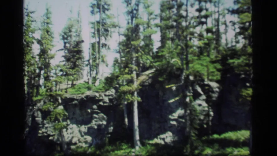 1977 Sapphire Lake, Montana 8mm frame showing a wider forested rock face with tall pines and scattered cut branches.