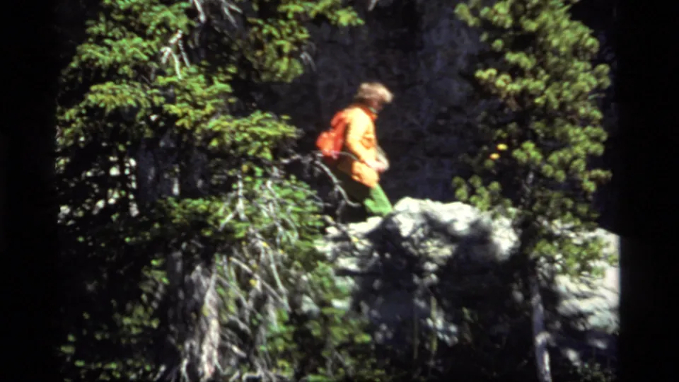 A backpacked hiker in an orange jacket crosses gray rocks in a forested Sapphire Lake, Montana home-movie scene.