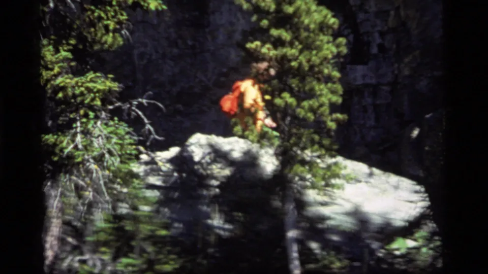 A hiker in orange stands on pale rocks amid dense evergreens in 1977 Sapphire Lake, Montana 8mm footage.