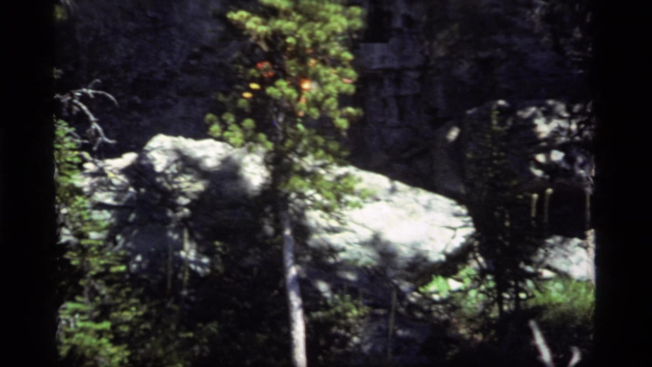 1977 Sapphire Lake, Montana home-movie frame showing pale boulders, evergreen growth, grass, and a dark rock face before the hiker enters view.