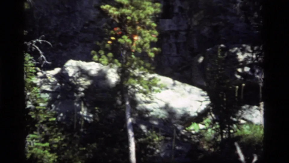 1977 Sapphire Lake, Montana home-movie frame showing pale boulders, evergreen growth, grass, and a dark rock face before the hiker enters view.