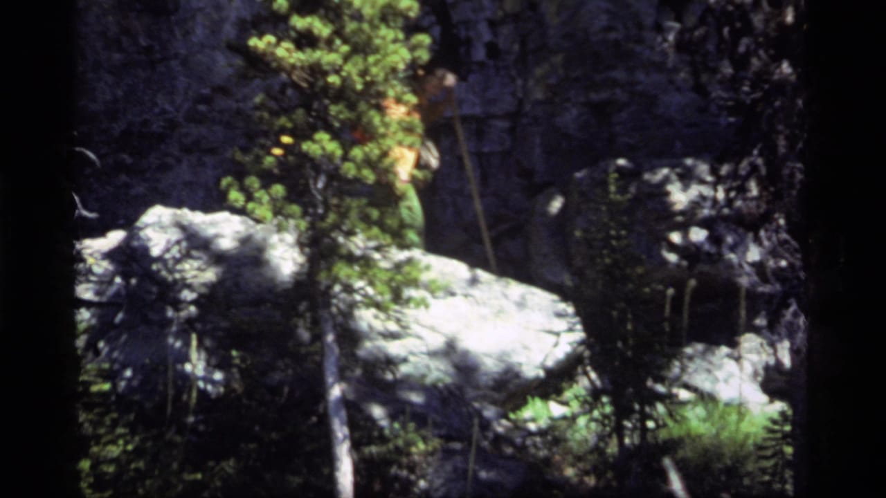 1977 Sapphire Lake, Montana frame with a hiker partly visible in an orange shirt behind evergreen foliage and pale boulders.