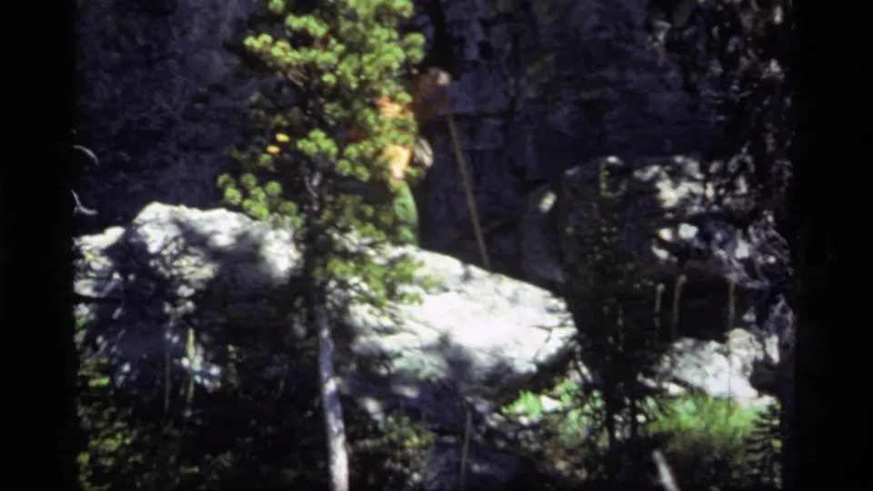 1977 Sapphire Lake, Montana frame with a hiker partly visible in an orange shirt behind evergreen foliage and pale boulders.