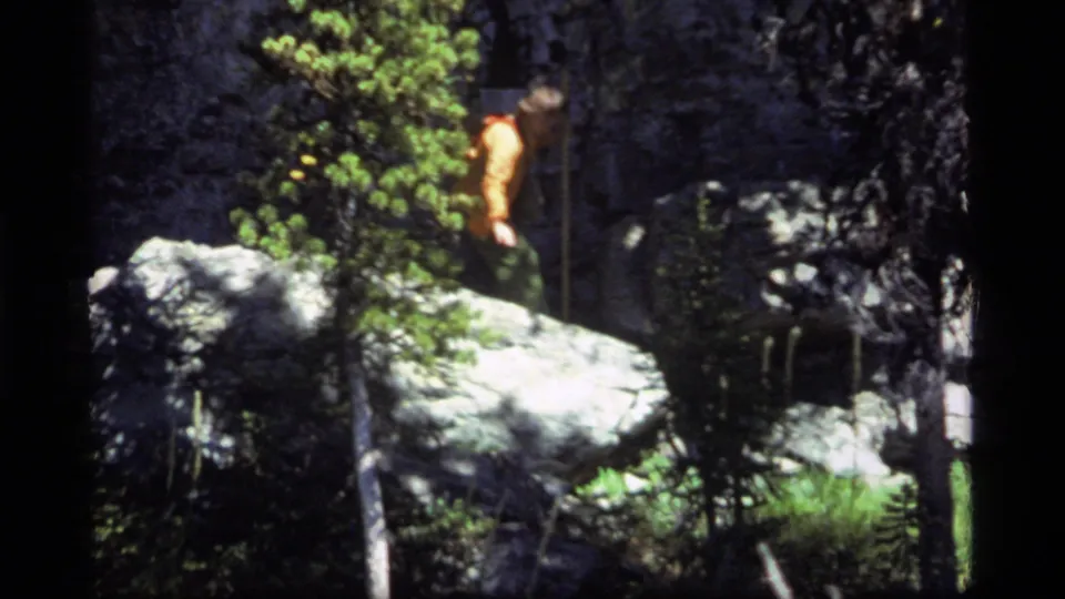 1977 Sapphire Lake, Montana frame showing a hiker in an orange shirt and green trousers moving carefully near large boulders.
