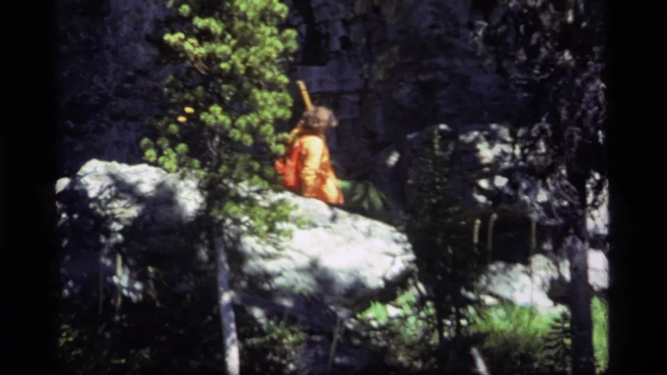 1977 Sapphire Lake, Montana frame showing the orange-shirted hiker descending rocky terrain with a walking stick among trees and boulders.