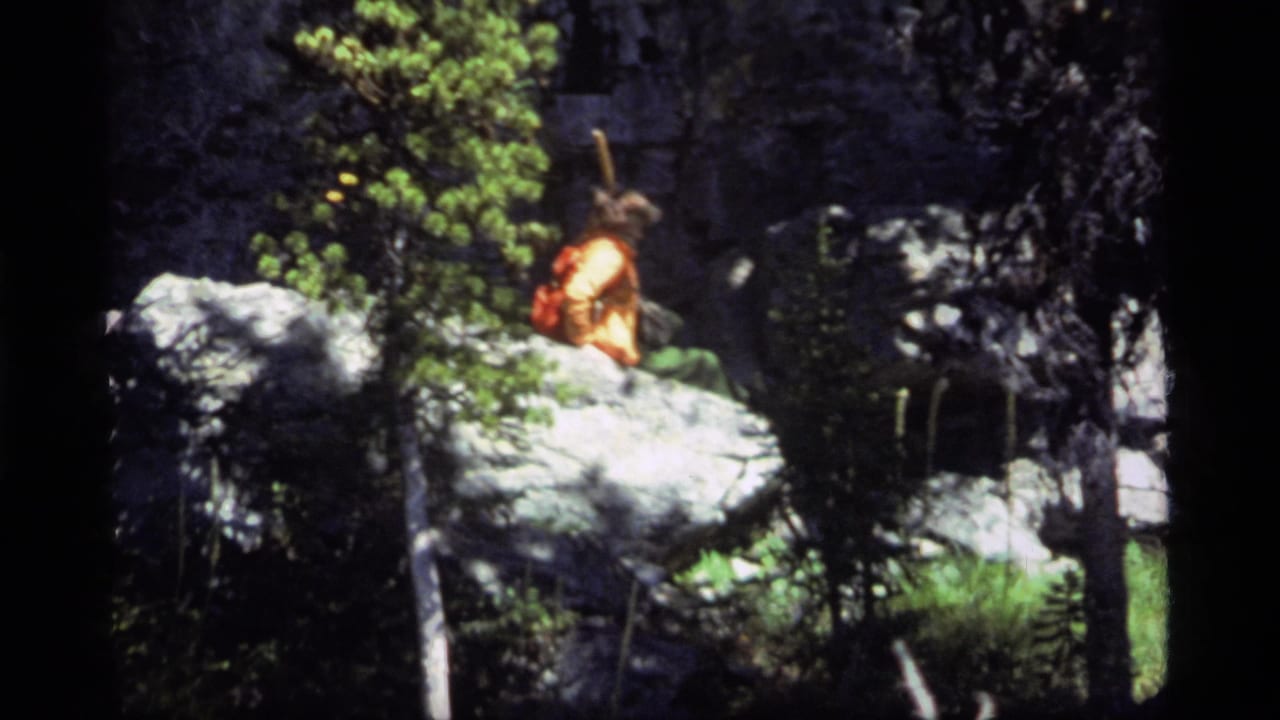 A hiker in orange outerwear with a red backpack appears on pale boulders in a shaded forest at Sapphire Lake, Montana, 1977.