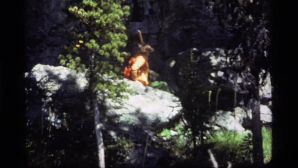 A hiker in orange outerwear with a red backpack appears on pale boulders in a shaded forest at Sapphire Lake, Montana, 1977.