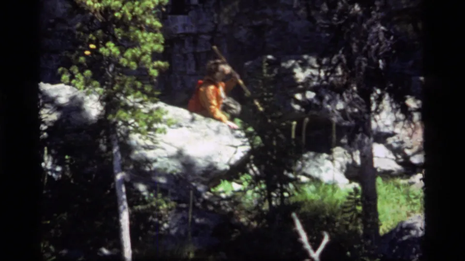 A backpacker in orange clothing is partly obscured by a bright rock ledge while moving through the shaded Sapphire Lake forest scene.