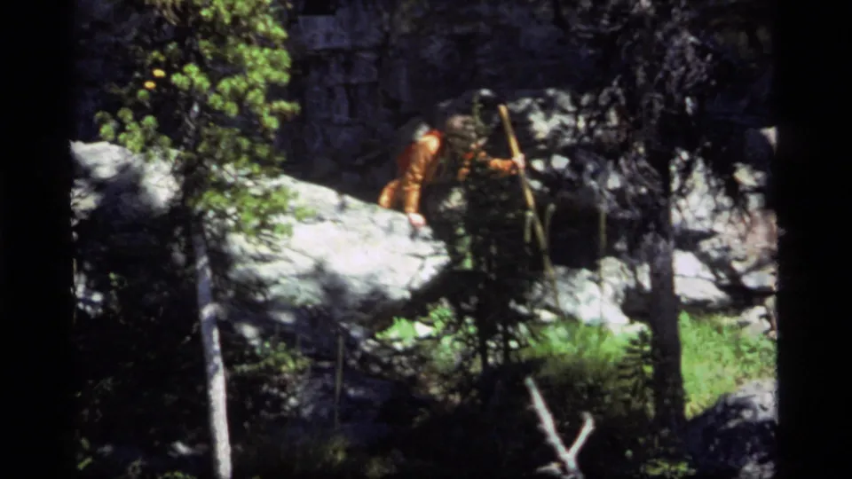 The hiker appears to use a walking stick or branch while stepping near boulders and trees in the 1977 Sapphire Lake home-movie footage.